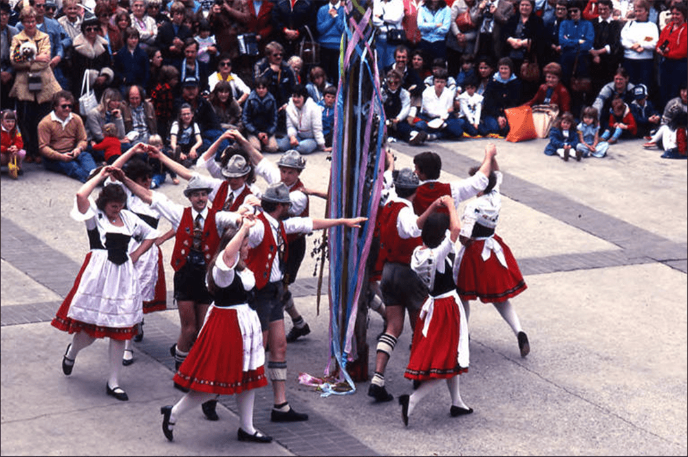 Crowd standing watching women in Bavarian dress dancing around a pole holding onto ribbons